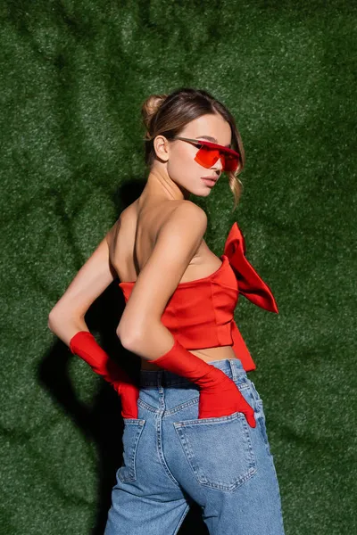 Стильна жінка в червоній блузці, сонцезахисних окулярах і рукавицях позує руками в кишенях джинсів біля трав'яного фону — стокове фото