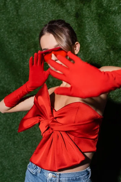 Femme élégante en chemisier rouge, lunettes de soleil et gants ne montrant aucun geste près de fond herbeux — Photo de stock