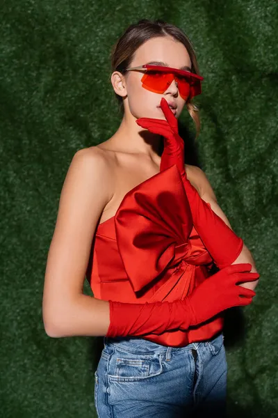 Modelo con estilo en blusa roja con lazo, gafas de sol y guantes posando cerca de fondo herboso - foto de stock