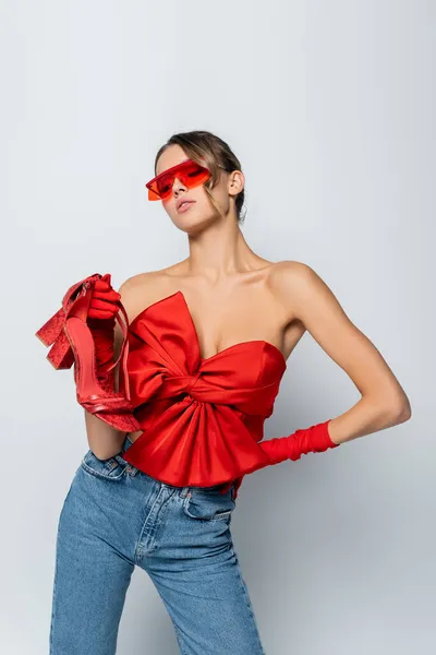 Modelo elegante en blusa roja, gafas de sol y guantes que sostienen los zapatos mientras posan con la mano en la cadera aislada en gris - foto de stock