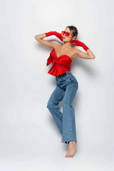 Full length of barefoot and stylish model in red blouse, sunglasses and gloves posing on grey — Stock Photo