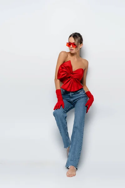 Full length of barefoot and trendy model in red blouse, sunglasses and gloves posing on grey — Stock Photo