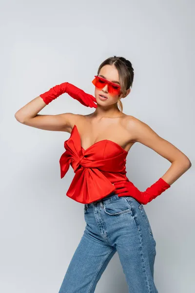 Стильна модель в червоній блузці, сонцезахисних окулярах і рукавичках, що стоять з рукою на стегні ізольовані на сірому — стокове фото