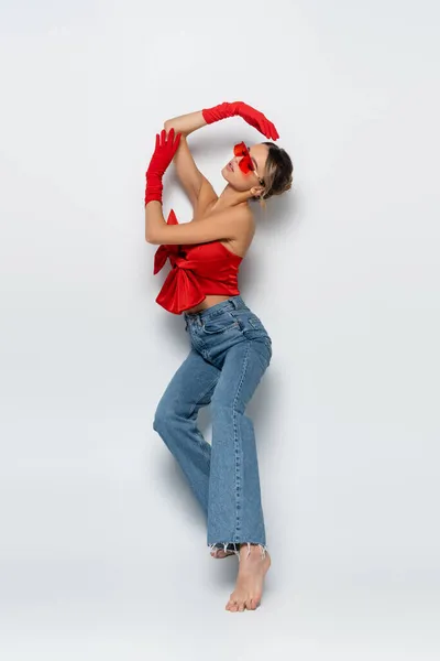Повна довжина босоніж молода жінка в червоній блузці, сонцезахисні окуляри і рукавички позують на сірому — Stock Photo