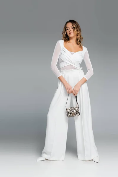 Full length of curly young woman in white outfit holding purse with zebra print and posing on grey — Stock Photo