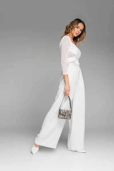 Full length of curly young model in white outfit holding purse with zebra print and posing on grey — Stock Photo