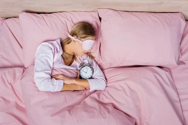 Top view of woman in sleep mask lying near alarm clock on pink bedding — Stock Photo