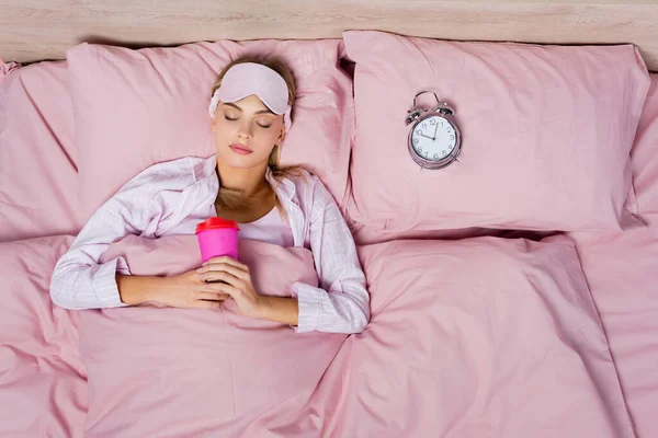 Vista superior de la mujer con la máscara del sueño sosteniendo taza de papel cerca del reloj despertador en la cama - foto de stock