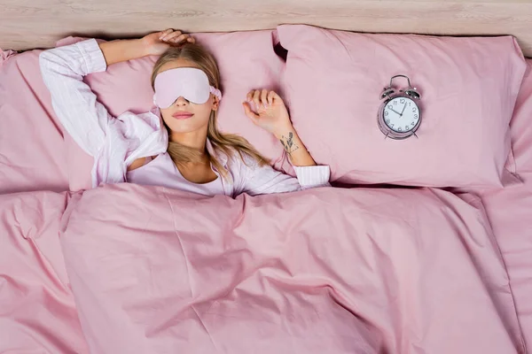 Draufsicht einer Frau in Schlafmaske und Schlafanzug, die neben dem Wecker auf dem Bett liegt — Stockfoto
