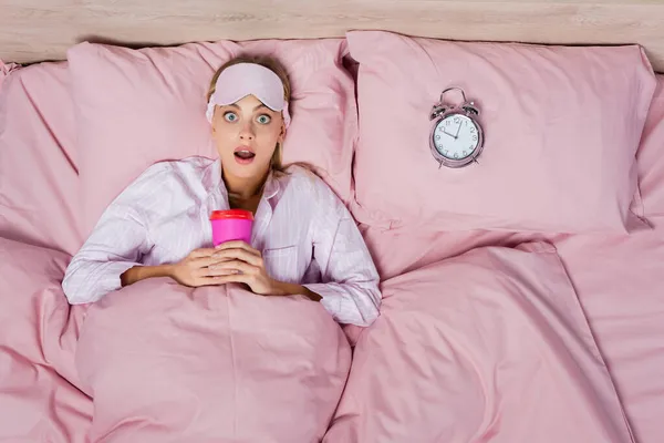 Вид сверху изумленной женщины в маске для сна, держащей бумажную чашку возле будильника на кровати — стоковое фото