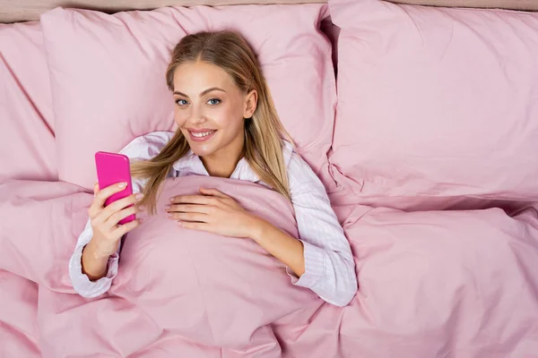 Top view of smiling woman using pink smartphone on bed — Stock Photo