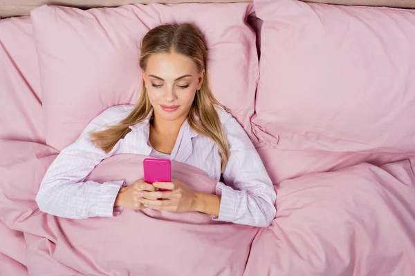 Вид сверху молодой женщины, пользующейся мобильным телефоном на розовом постельном белье — стоковое фото