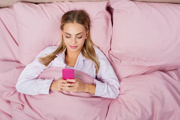 Vue du dessus de la jeune femme utilisant un téléphone portable sur la literie rose — Photo de stock