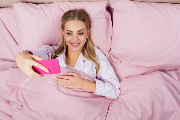 Vue du dessus de la femme blonde souriante prenant selfie sur smartphone sur literie rose — Photo de stock