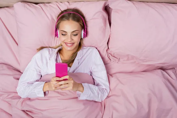 Visão superior da mulher alegre em fones de ouvido usando smartphone na cama rosa — Fotografia de Stock