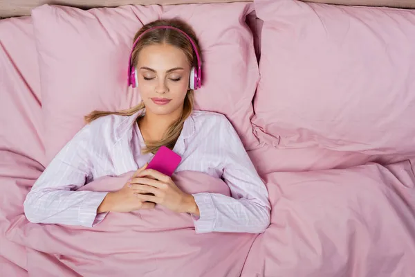 Top view of young woman in headphones holding smartphone and sleeping on bed — Stock Photo