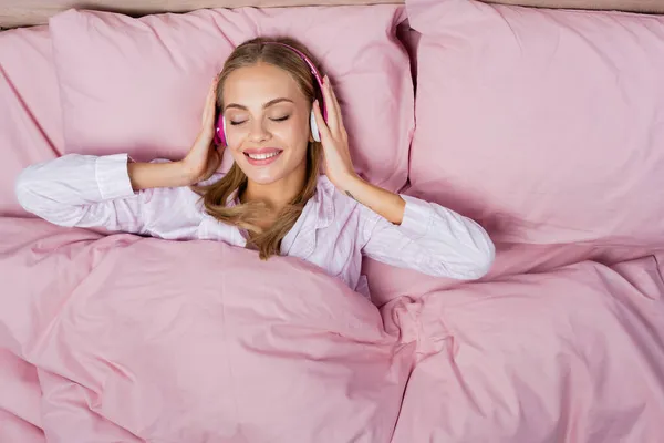 Draufsicht auf eine fröhliche Frau, die im Bett mit Kopfhörern Musik hört — Stockfoto