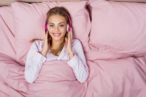 Draufsicht einer Frau mit Kopfhörern, die auf dem Bett in die Kamera lächelt — Stockfoto