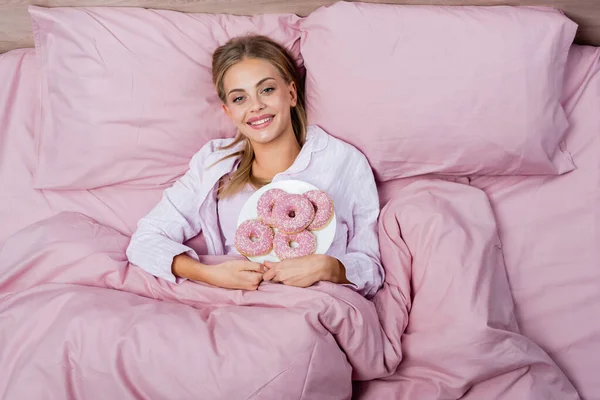 Vista dall'alto di allegra giovane donna che tiene il piatto con ciambelle sul letto — Foto stock