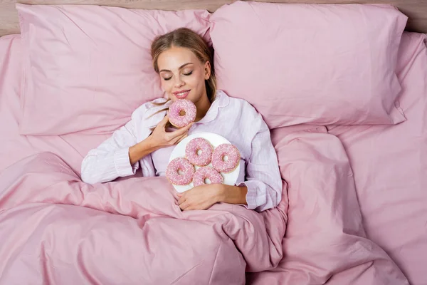 Vue du dessus de la femme souriante tenant beignet sur lit rose à la maison — Photo de stock