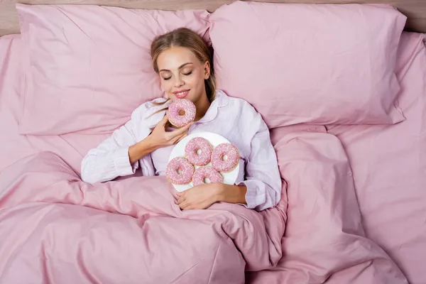 Top view of smiling woman holding donut on pink bed at home — Stock Photo