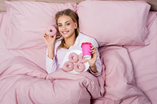 Vue du dessus de la femme souriante tenant beignet et tasse en papier sur le lit à la maison — Photo de stock