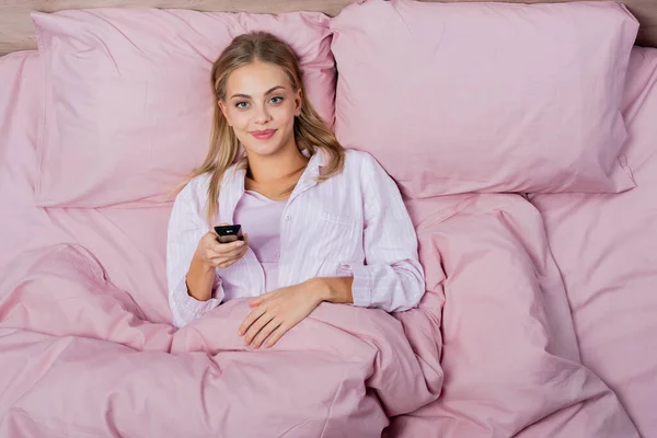 Vista dall'alto della donna bionda sorridente che tiene il telecomando sul letto rosa — Foto stock