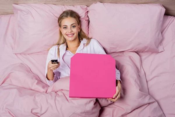 Visão superior da mulher sorridente no pijama segurando controle remoto e caixa de pizza na cama — Fotografia de Stock