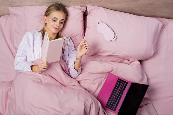 Vue du dessus de la femme souriante tenant le cahier et le stylo près de l'ordinateur portable et le masque de sommeil sur le lit — Photo de stock