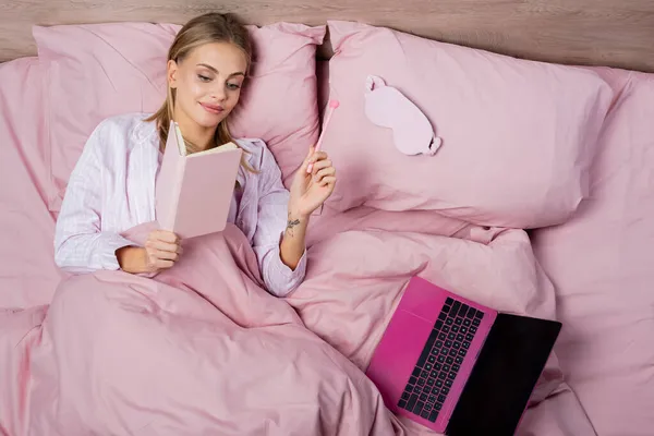 Vista superior de la mujer sonriente sosteniendo portátil y pluma cerca de la computadora portátil y la máscara del sueño en la cama - foto de stock