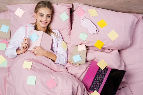 Top view of positive woman holding pen and notebook near books, laptop and sticky notes on bed — Stock Photo