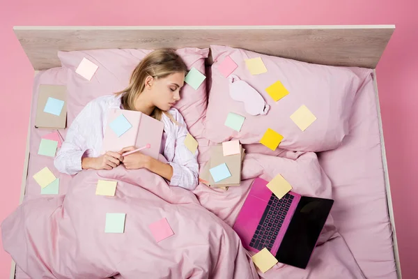 Вид сверху женщины с ноутбуком, спящей рядом с ноутбуком, книгами и записками на кровати на розовом фоне — стоковое фото