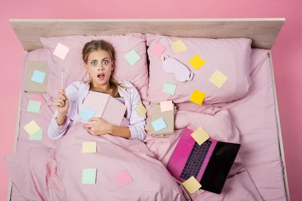 Vista dall'alto della donna scioccata che tiene il notebook vicino a libri, laptop e appunti appiccicosi sul letto su sfondo rosa — Foto stock