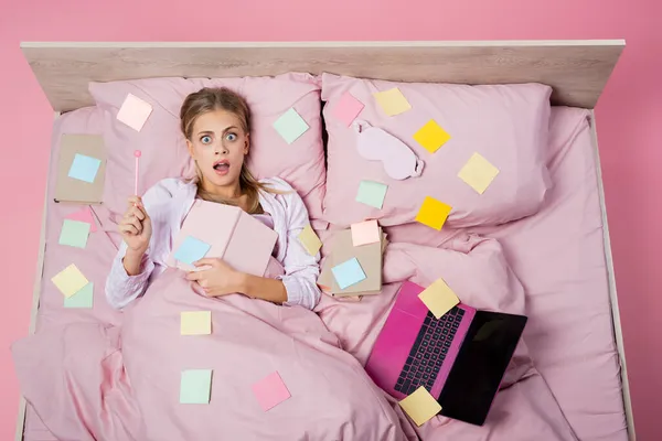 Top view of shocked woman holding notebook near books, laptop and sticky notes on bed on pink background — Stock Photo