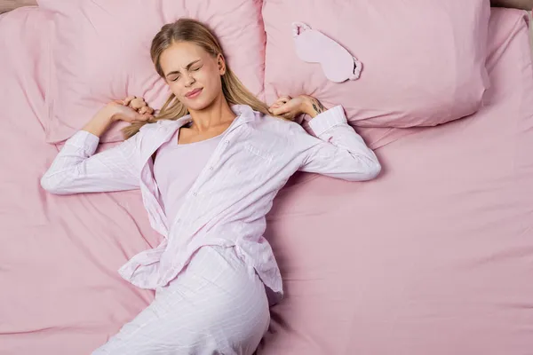 Draufsicht einer jungen Frau im Schlafanzug, die sich in der Nähe einer Schlafmaske auf dem Bett ausbreitet — Stockfoto
