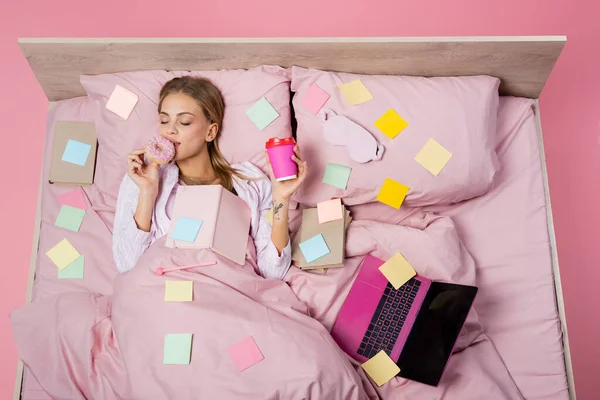 Vista dall'alto della donna che tiene il caffè per andare e ciambella vicino a libri, laptop e appunti appiccicosi sul letto su sfondo rosa — Foto stock