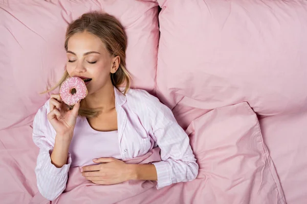 Top view of smiling woman in pajama holding donut on pink bed — Stock Photo