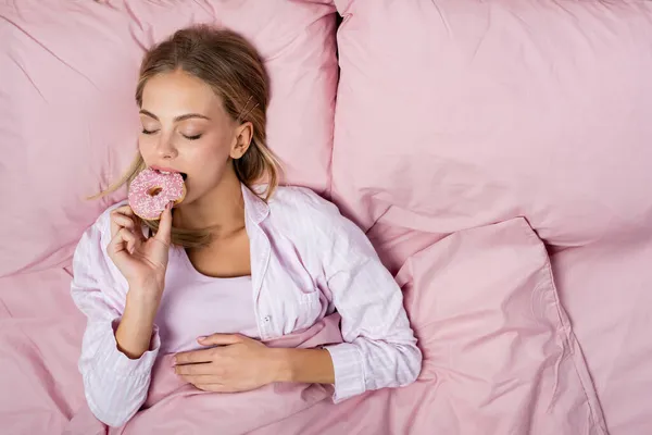 Draufsicht einer Frau im Pyjama, die Donut auf dem Bett isst — Stockfoto