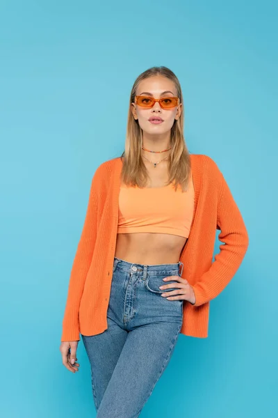 Mujer rubia en gafas de sol cogida de la mano en la cadera aislada en azul - foto de stock