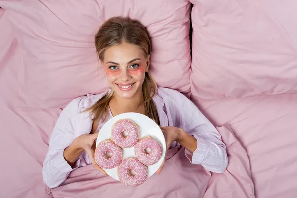 Top view of smiling woman with eye patches holding plate with donuts on bed — Stock Photo