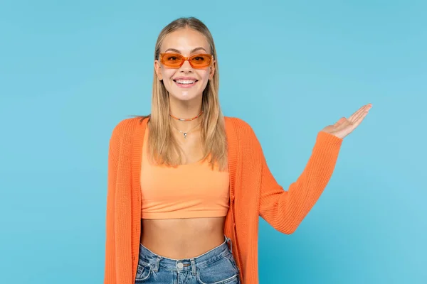 Mujer sonriente en gafas de sol naranjas apuntando con la mano aislada en azul — Stock Photo