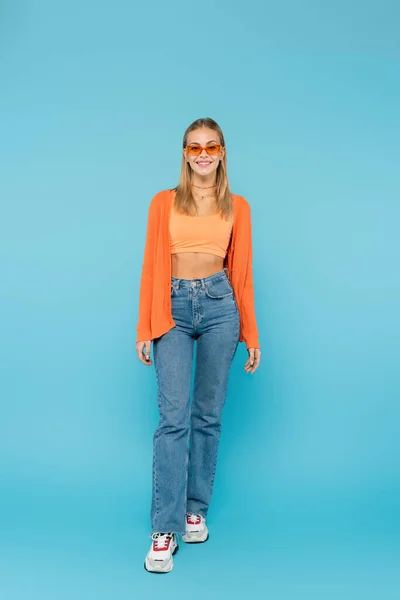 Full length of smiling woman in sunglasses and jeans looking at camera on blue background — Stock Photo