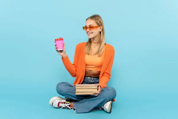 Estudiante positivo en gafas de sol sosteniendo café para llevar y libros sobre fondo azul - foto de stock