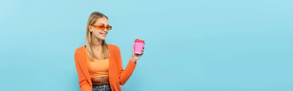 Femme blonde joyeuse dans des lunettes de soleil tenant café pour aller isolé sur bleu, bannière — Photo de stock