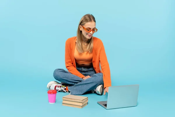 Positive student in sunglasses using laptop near books and paper cups on blue background — Stock Photo