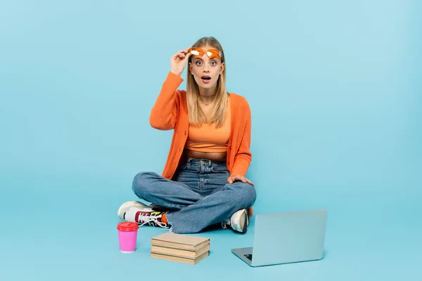 Studente stupito che tiene gli occhiali da sole vicino al computer portatile, caffè da andare e libri su sfondo blu — Foto stock