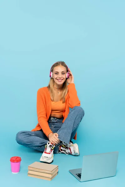 Happy blonde student in headphones sitting near laptop, paper cup and books on blue background — Stock Photo