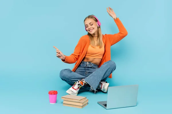 Étudiant blond joyeux dans des écouteurs assis près d'un ordinateur portable, des livres et du café pour aller sur fond bleu — Photo de stock