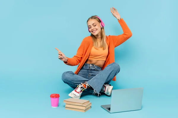 Cheerful blonde student in headphones sitting near laptop, books and coffee to go on blue background — Stock Photo