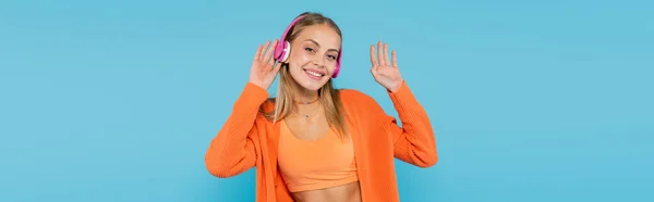 Femme blonde souriante dans les écouteurs agitant la main à la caméra isolée sur bleu, bannière — Photo de stock